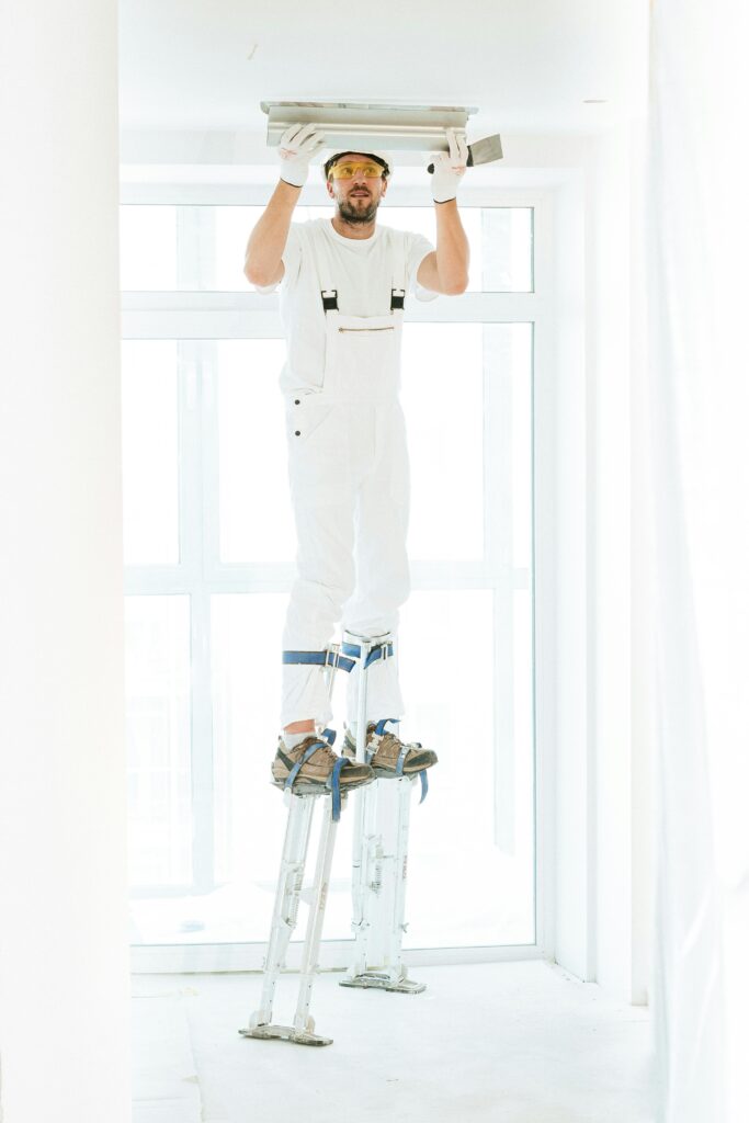 A construction worker in white jumpsuit on stilts plastering a ceiling indoors.